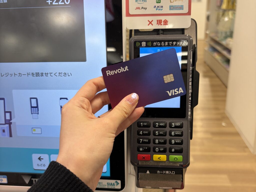 A person making a contactless payment at a store counter in Japan using a Revolut card held over a card reader terminal.